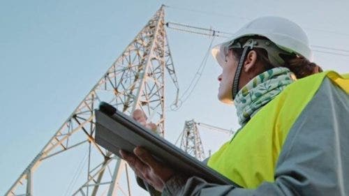 An electrical engineer makes a note on a tablet while looking up at a transmission tower.